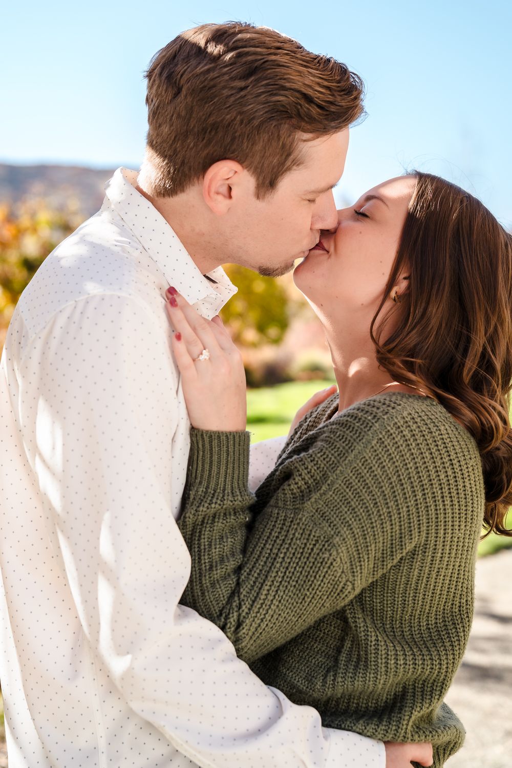A romantic couple embraces and kisses outdoors in autumn wearing a white shirt and olive green sweater.