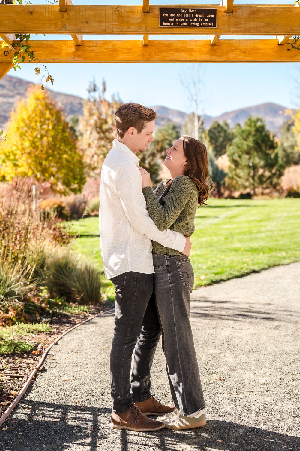 A couple embraces on a gravel path surrounded by autumn foliage and mountain scenery in the background.