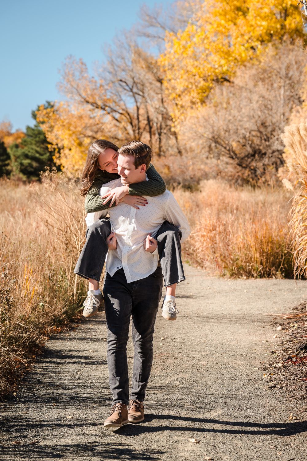 Two people share a playful moment together on a dirt path surrounded by golden autumn foliage and tall grass.