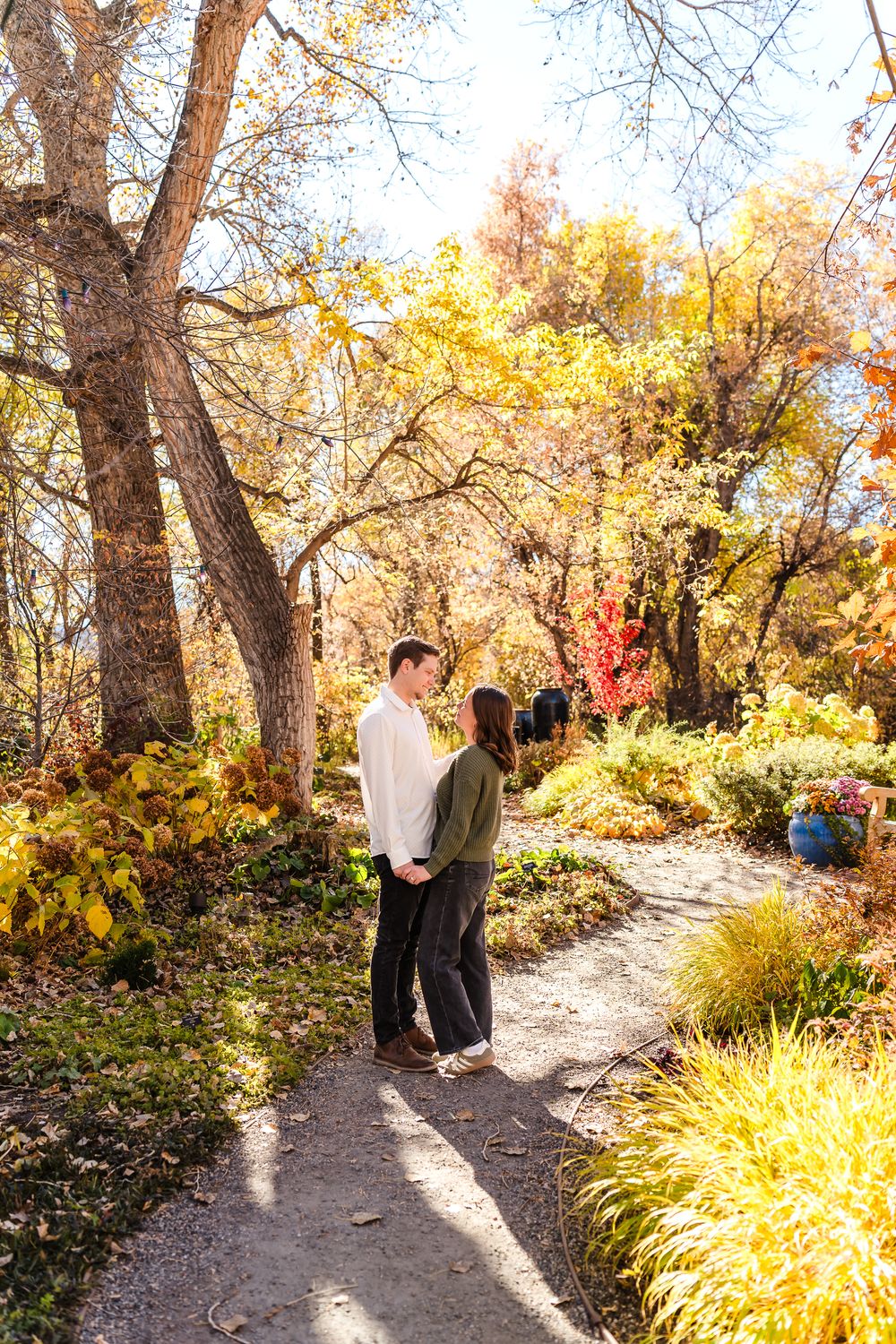 A couple stands together on a sunlit autumn path surrounded by golden foliage and ornamental grasses.