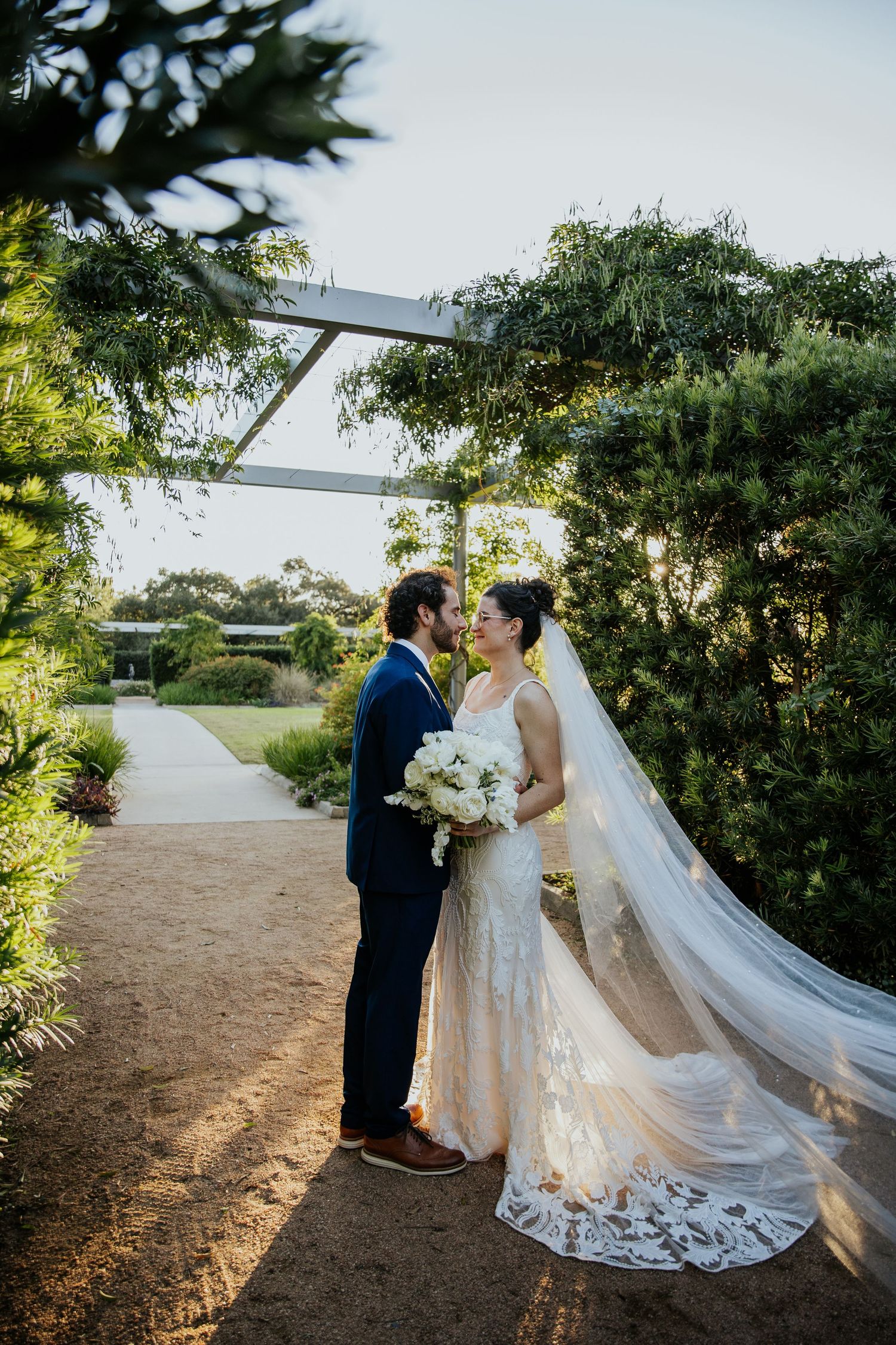 Bride and groom pose together on garden path with flowing white wedding dress and veil.