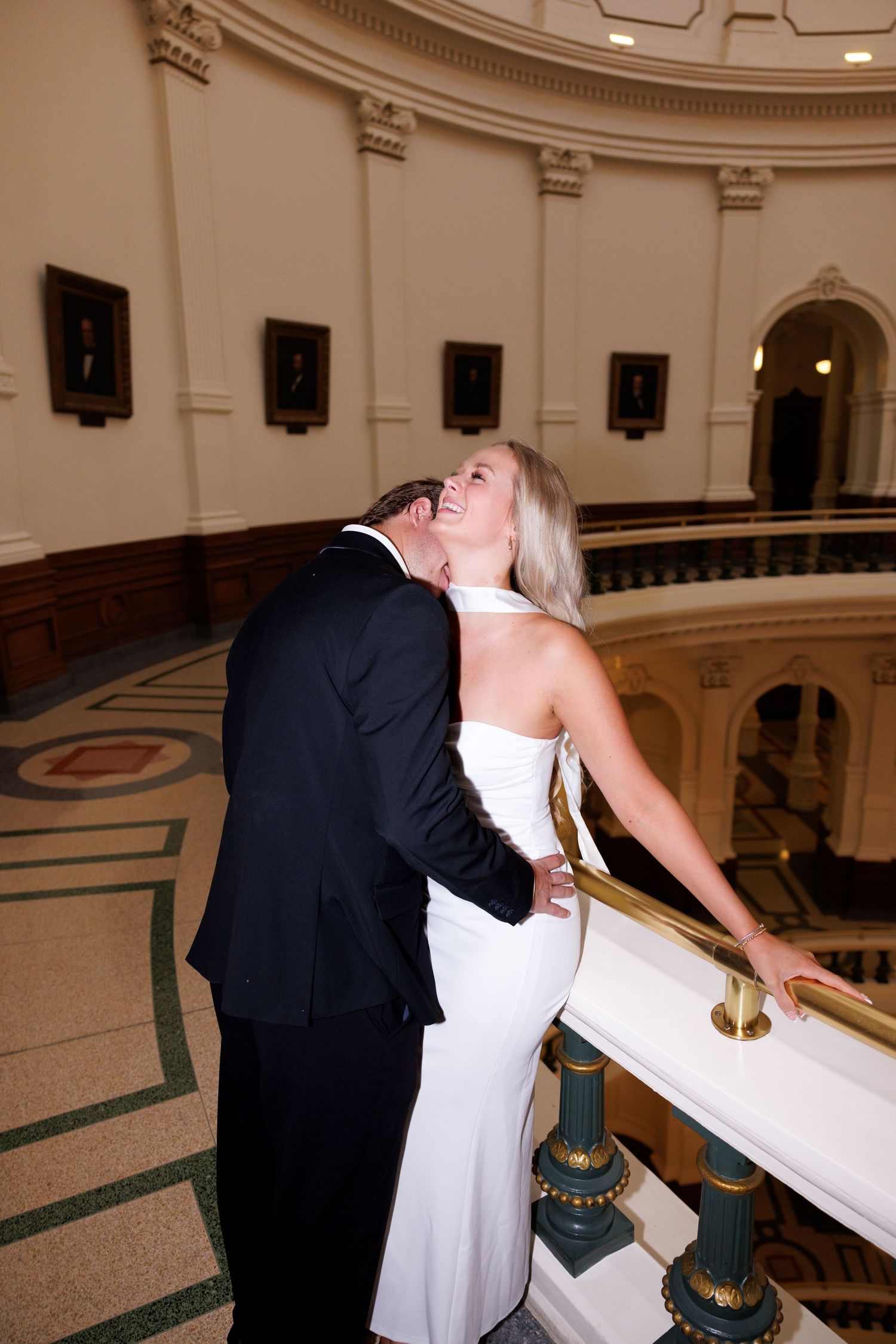 A couple shares a romantic moment along an ornate marble railing in a grand rotunda.