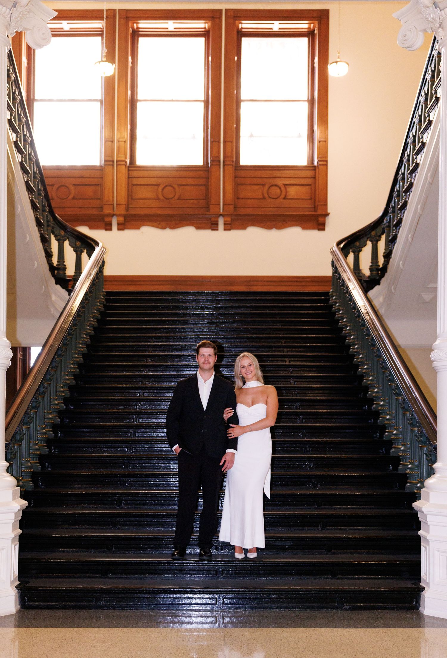 Wedding couple stands at bottom of grand staircase with ornate wooden details.