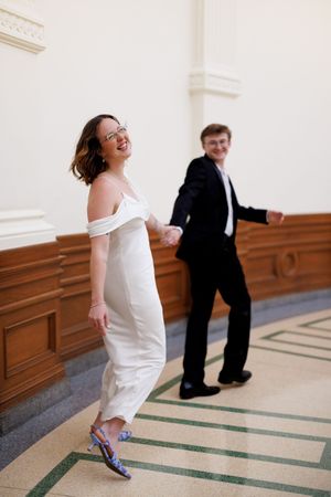 A playful couple in formal attire dances in a grand hallway with decorative floor patterns.