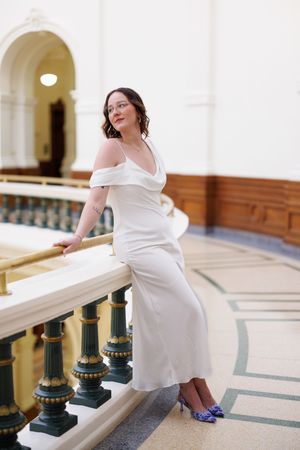 A person in a white dress sits elegantly on a marble railing with ornate green columns in a historic building.