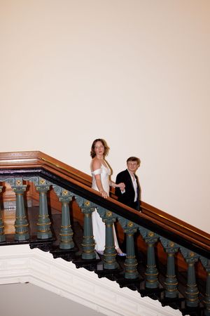 A couple poses on an ornate staircase with decorative balusters and white walls.