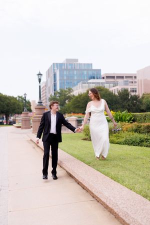 A couple in formal attire walks together on a path with city buildings in the background.