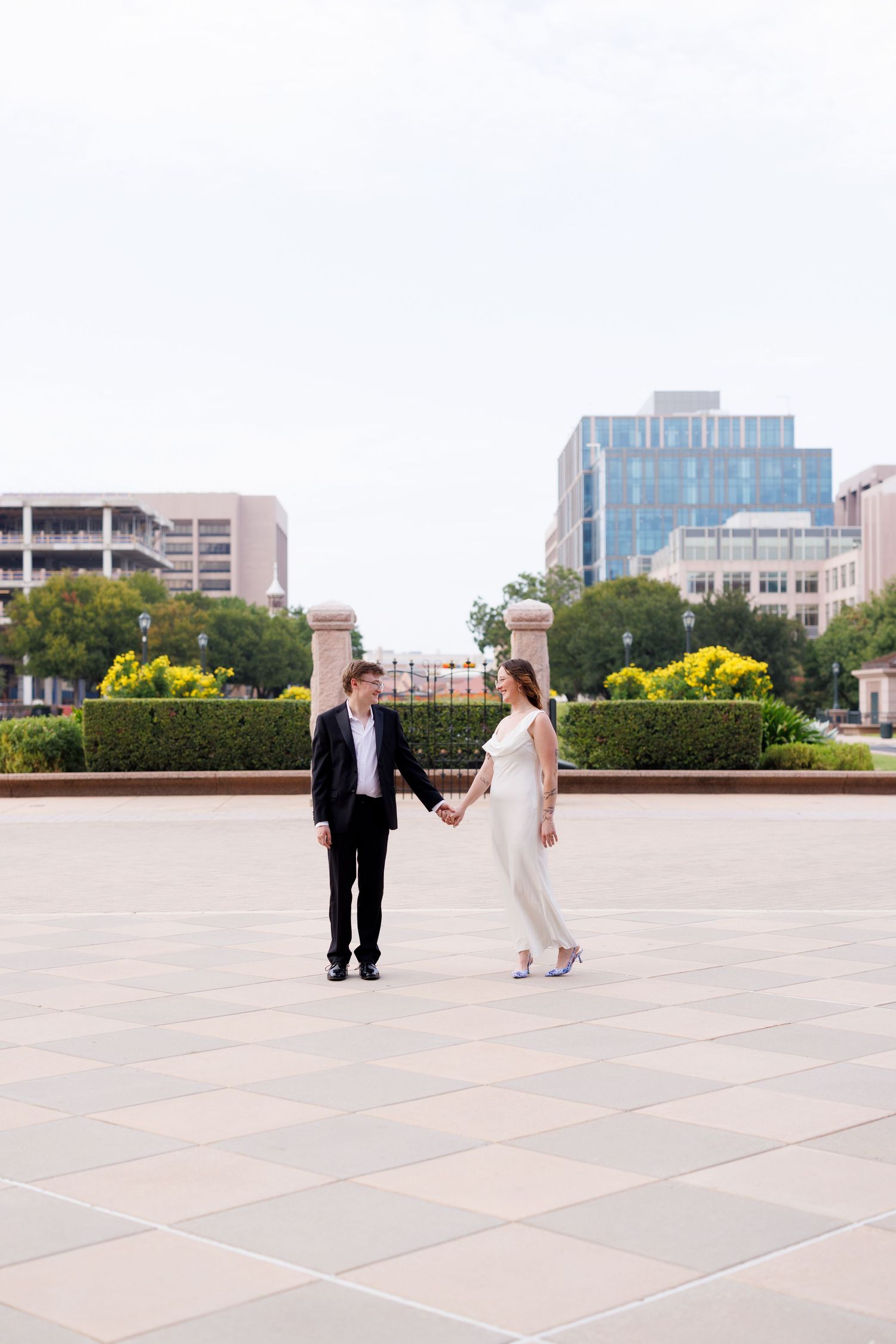 A couple dances playfully on an outdoor plaza with urban architecture in the background.