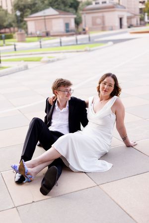 A couple in wedding attire sits casually on stone pavement during a photo session.