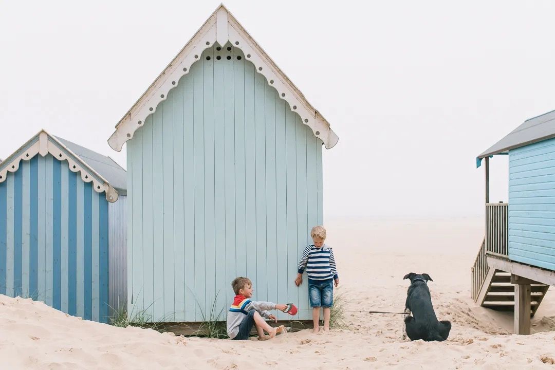 Two children and a dog sitting outside pastel blue beach huts on a sandy beach.