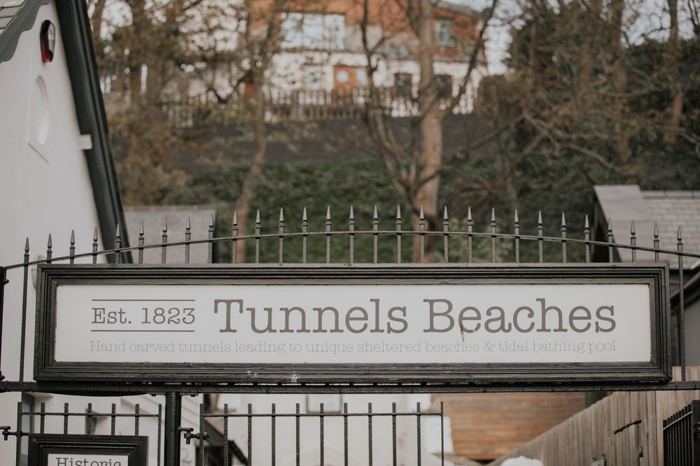 Vintage sign reading Est. 1823 Tunnels Beaches mounted on black iron fence with trees behind.