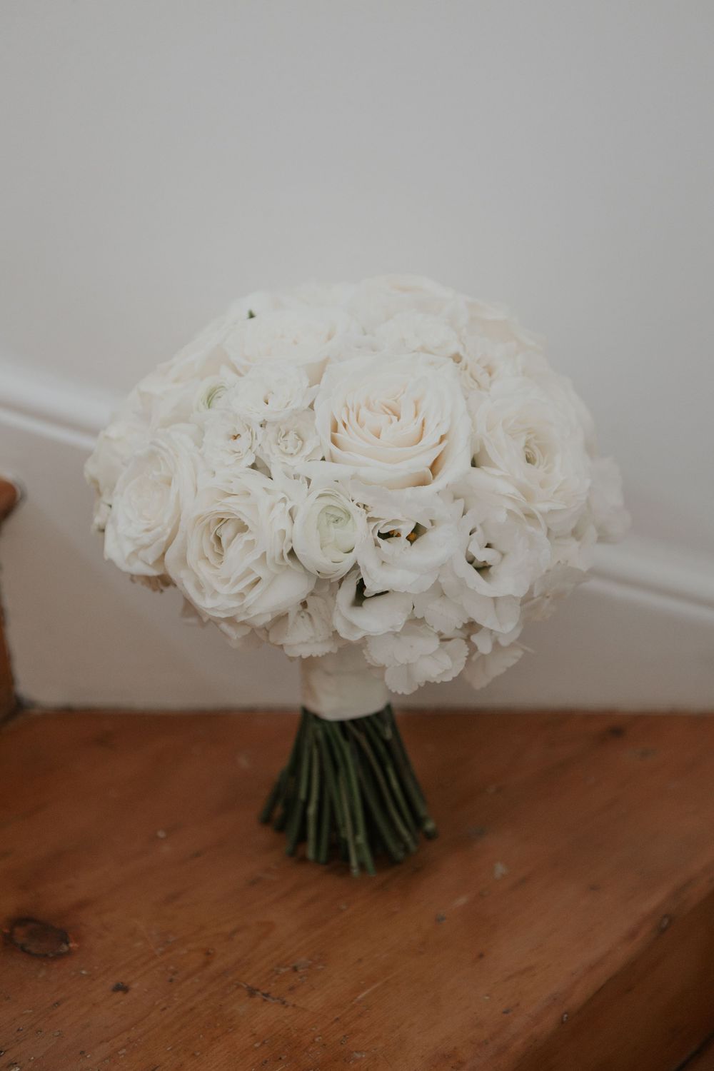 Classic round bridal bouquet of white roses and ranunculus tied with green stems on wood surface.