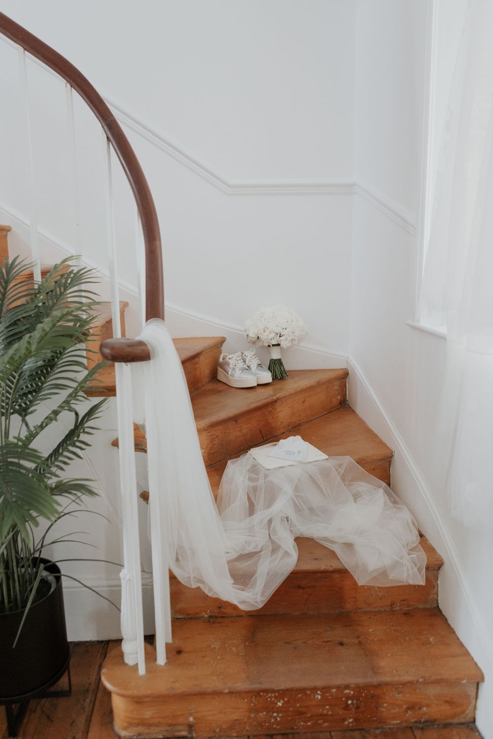 Bridal veil draped over wooden staircase with white floral bouquet and green plant in elegant home.