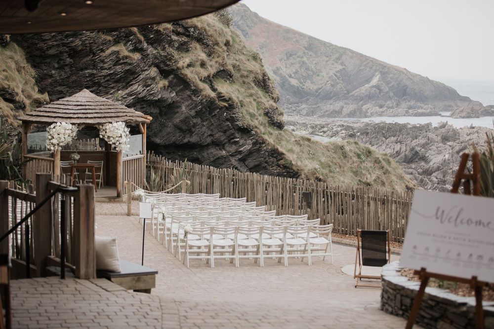 Outdoor beach wedding ceremony setup with white chairs, wooden gazebo, and dramatic coastal cliffs.
