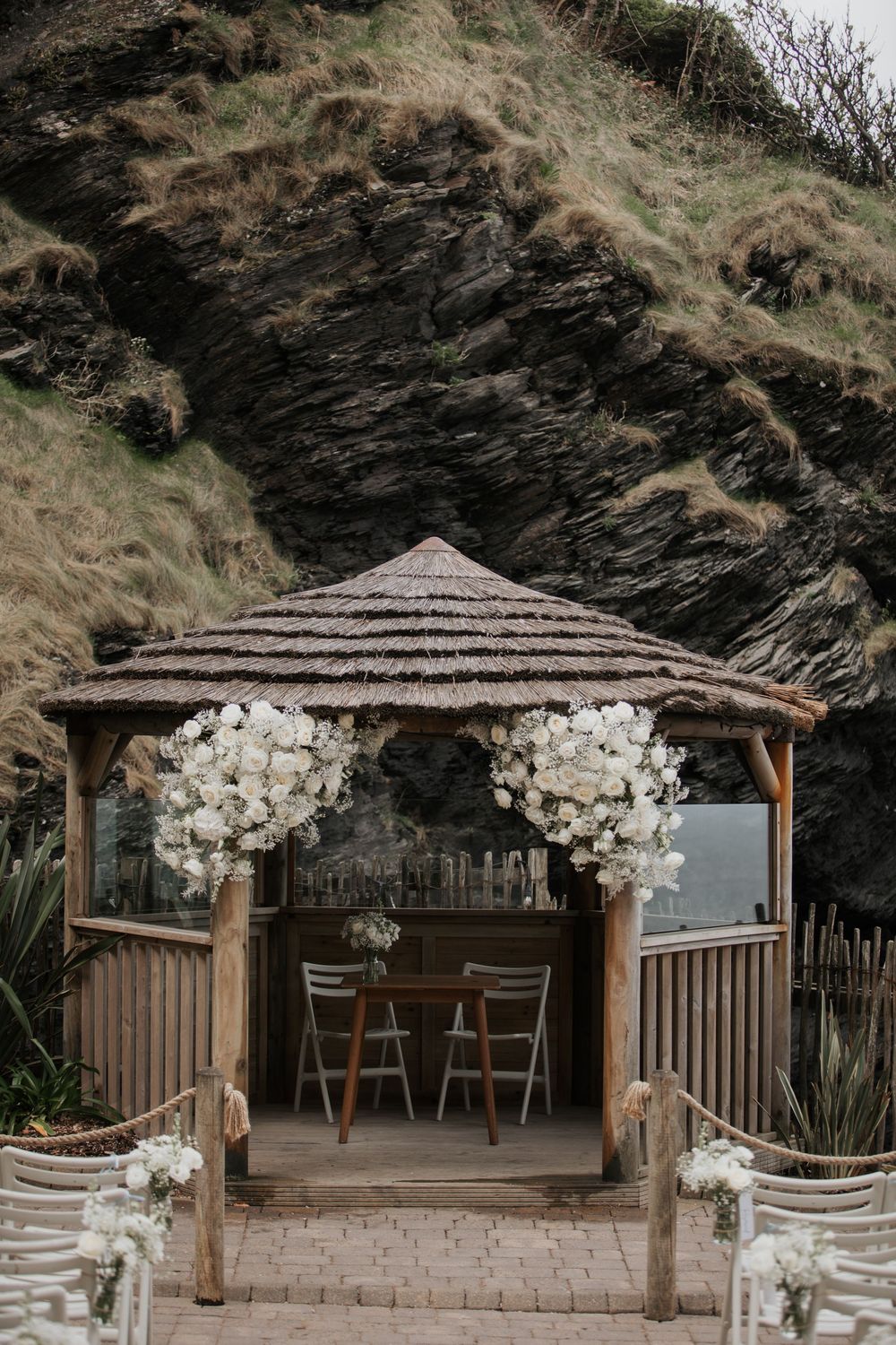 Rustic wooden wedding gazebo adorned with large white floral arrangements against rocky coastal cliffs.