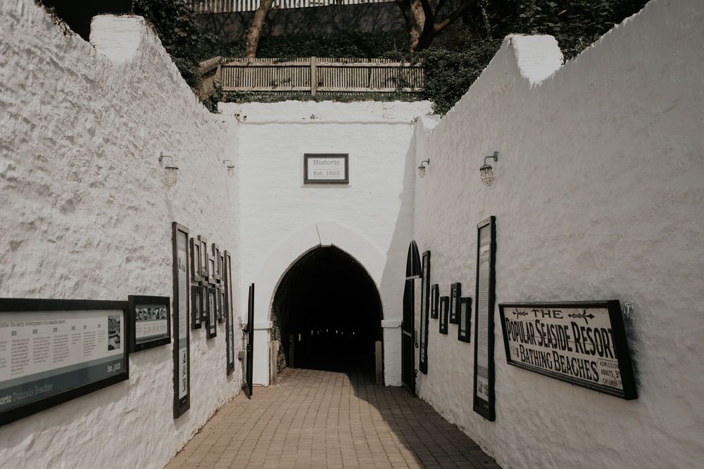 White-walled tunnel entrance with arched doorway and framed artwork leading to a dark passageway.
