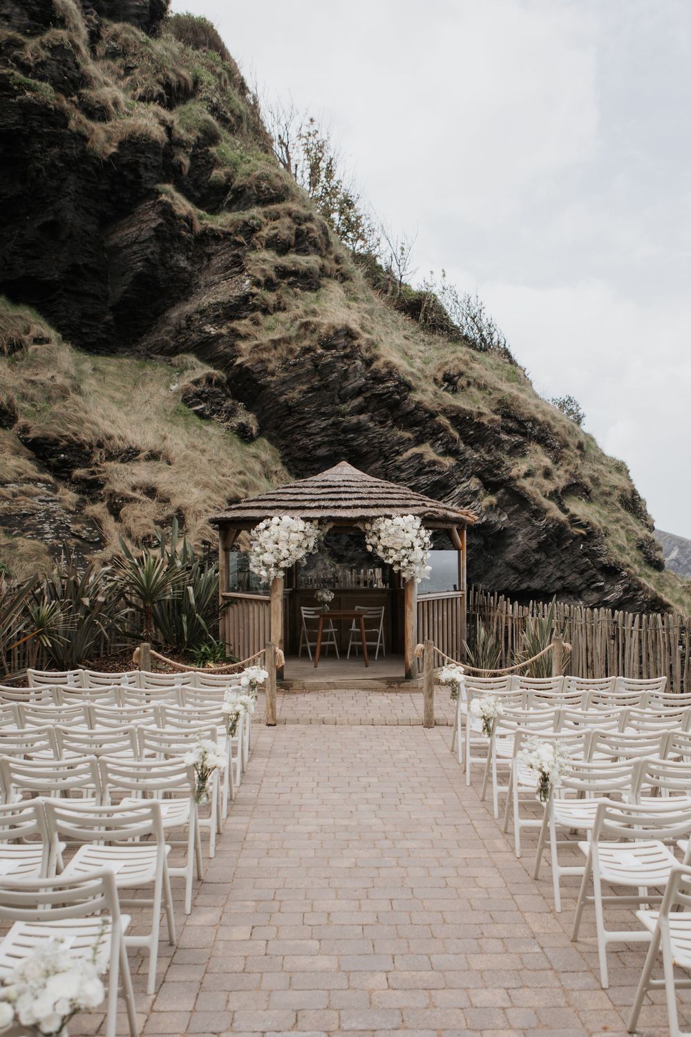 Elegant outdoor coastal wedding ceremony with white chairs, floral gazebo altar, and green clifftop backdrop.