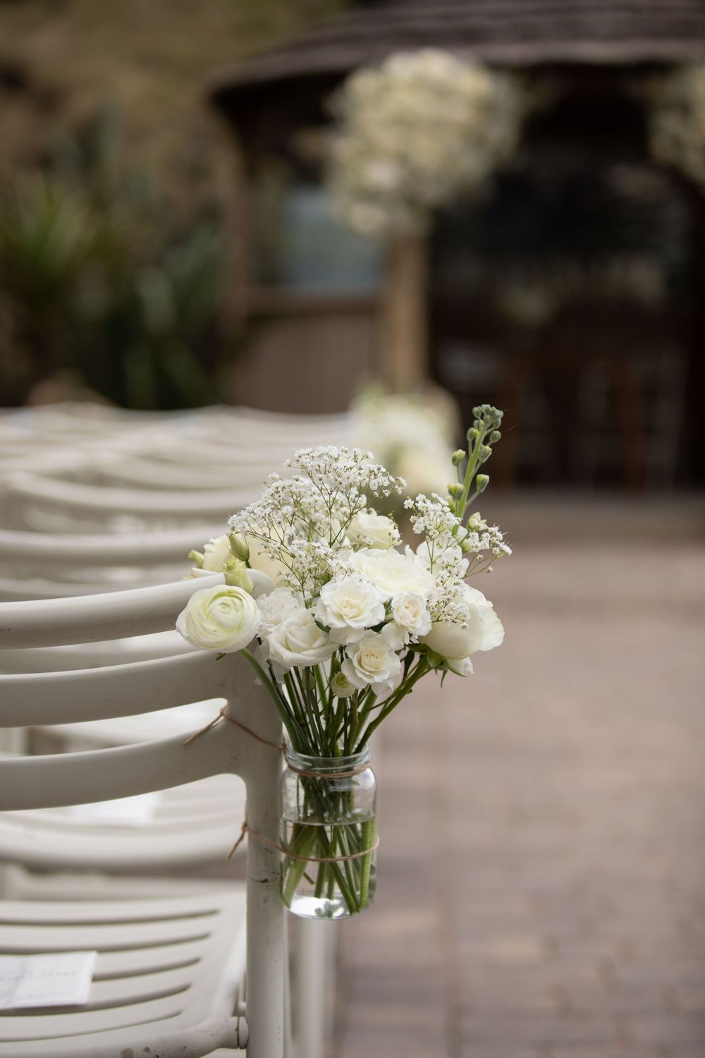 White roses and baby's breath in glass jar vase decorating wedding ceremony aisle chairs outdoors.