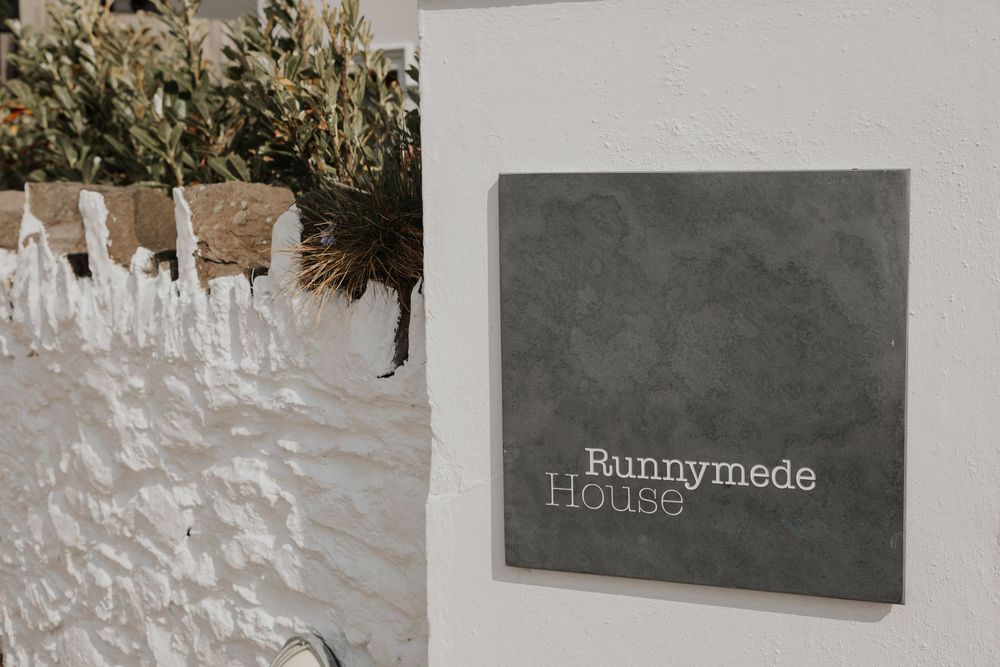 Grey slate sign reading Runnymede House mounted on a whitewashed stone wall with dried flowers.