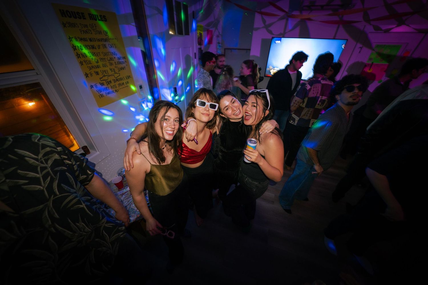 Group of four young women posing together at a colorful indoor party with disco lights.