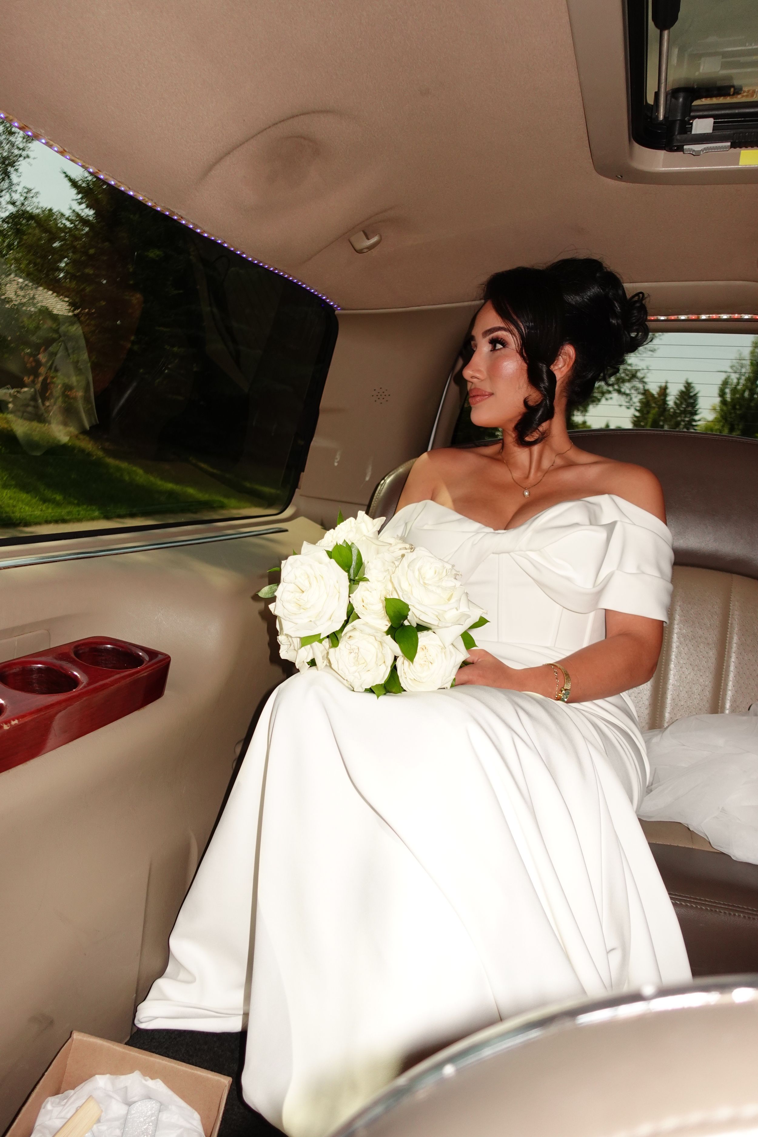 A bride in a white wedding dress sits in the backseat of a car holding a bouquet of white roses.