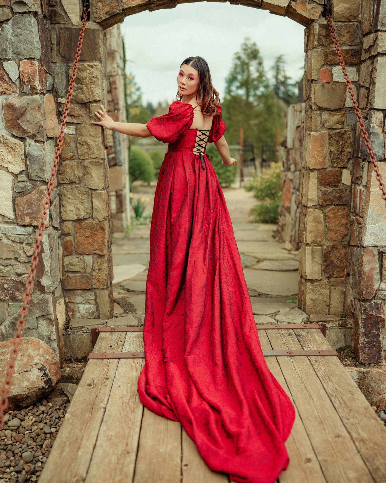 A stunning red ball gown with a long train photographed in a rustic stone archway with a wooden walkway.