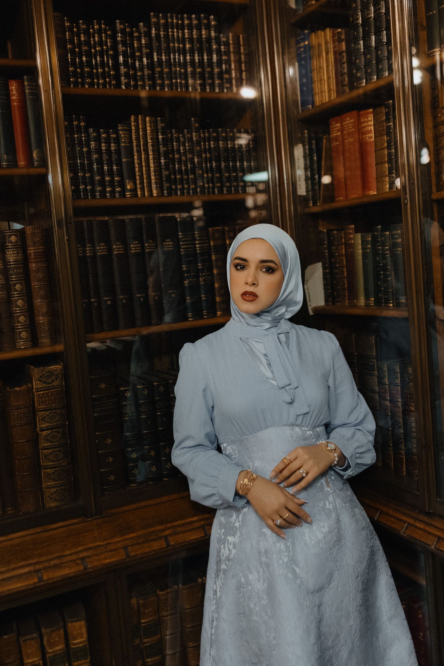 A person in a light blue dress and hijab stands in a vintage library surrounded by antique leather-bound books.