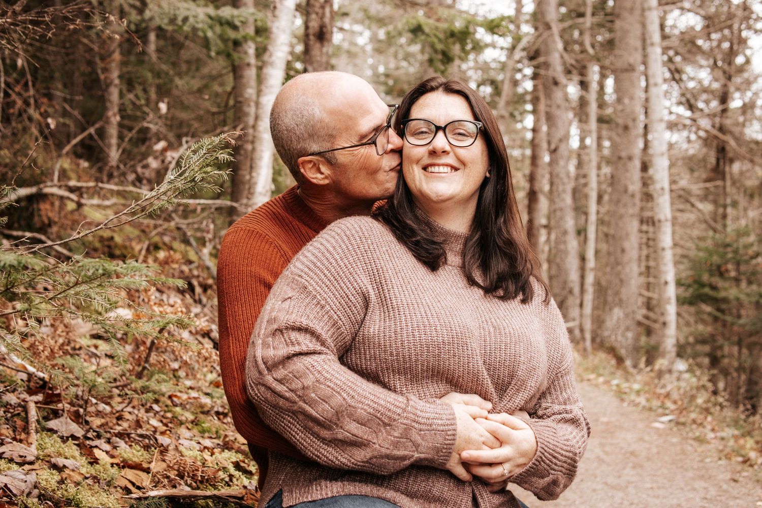 A couple in cozy sweaters share tender moments along a wooded forest path during an autumn engagement photoshoot.
