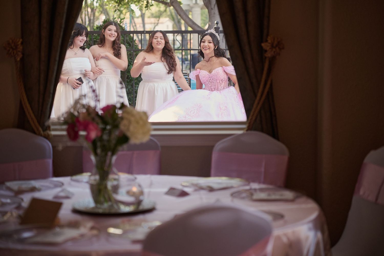A wedding party peeks through a window at an elegant reception table with pink linens and floral centerpiece.