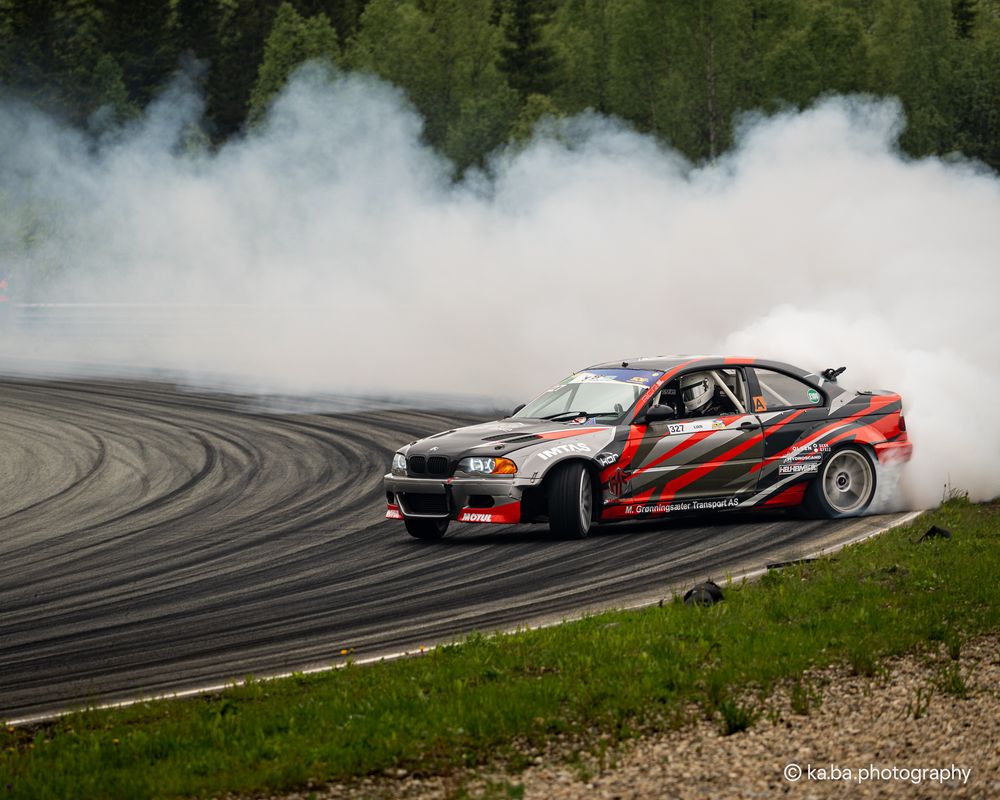 Red and black BMW drifting on a race track surrounded by smoke and forest.