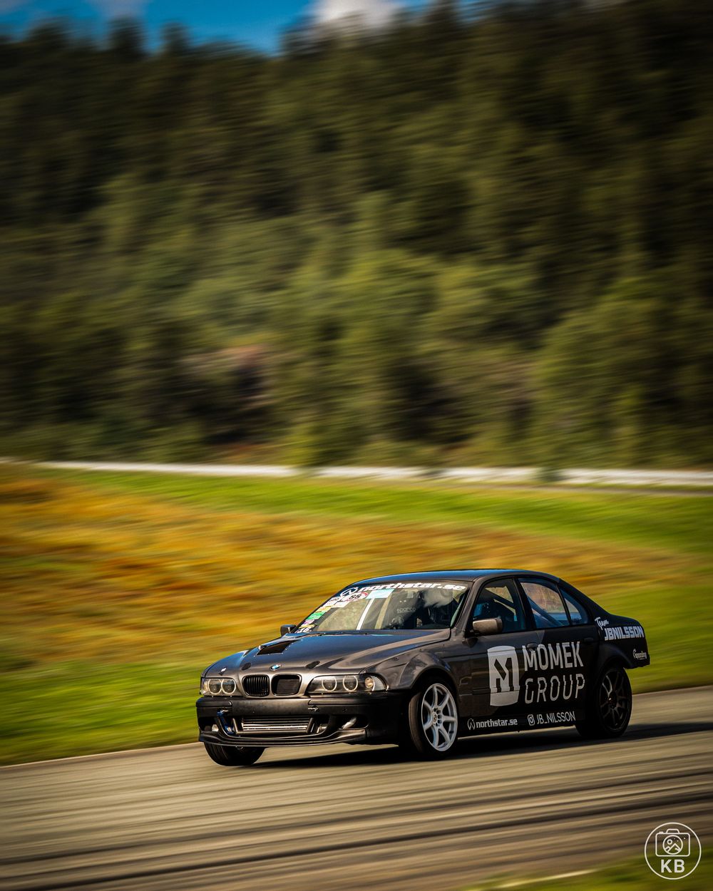 Gray BMW sedan with Momek Group livery racing on a track surrounded by green forest.