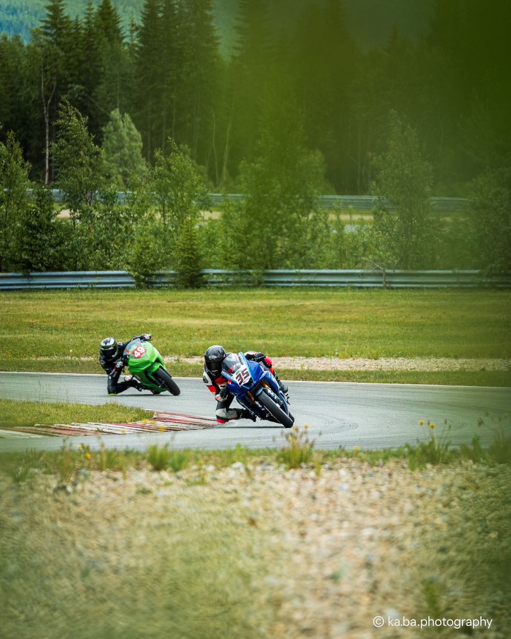 Two motorcycle racers compete on a scenic race track surrounded by lush green forest.
