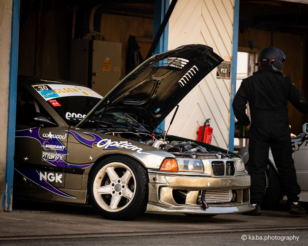 Modified BMW race car with hood open displaying sponsor decals parked in a garage bay.