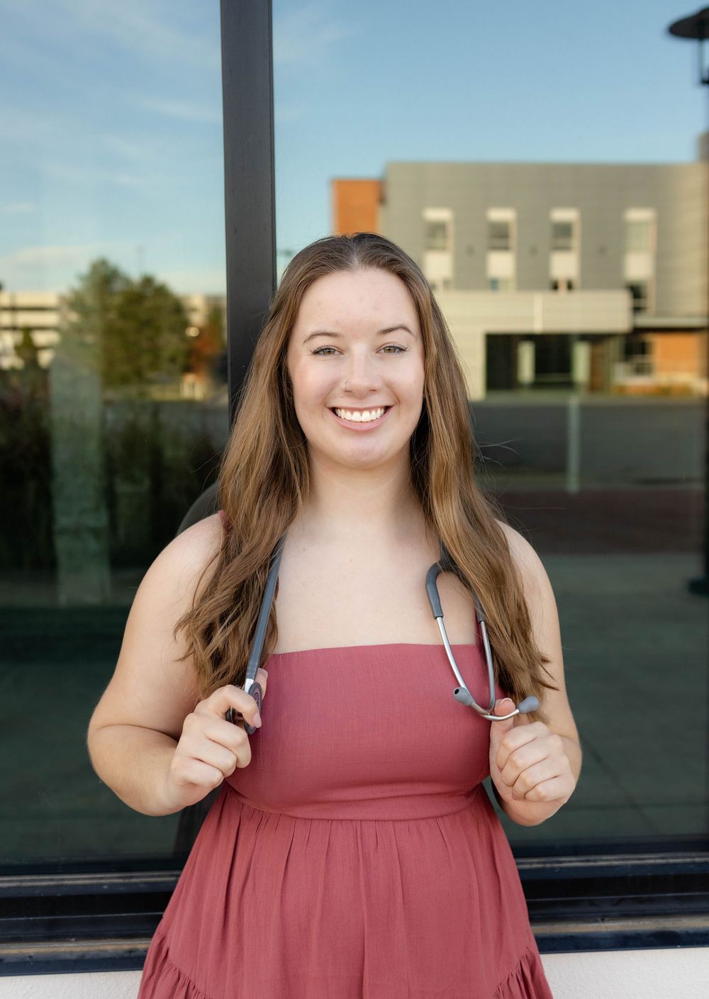 True West Photography - Healthcare student in burgundy dress poses with stethoscope outside medical building.