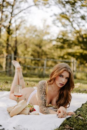 Romantic outdoor boudoir photo with model lounging on white blanket with wine.