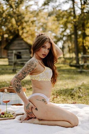 Artistic boudoir photoshoot in a sun-dappled outdoor setting with rustic cabin and autumn foliage in background.