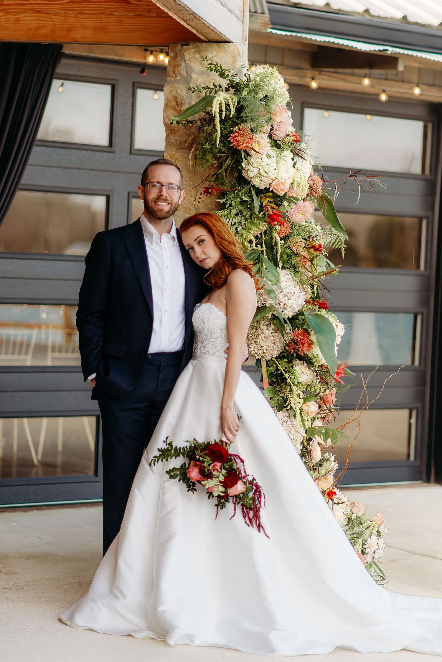 A romantic wedding couple poses together in front of a modern industrial venue with elegant floral arrangements.