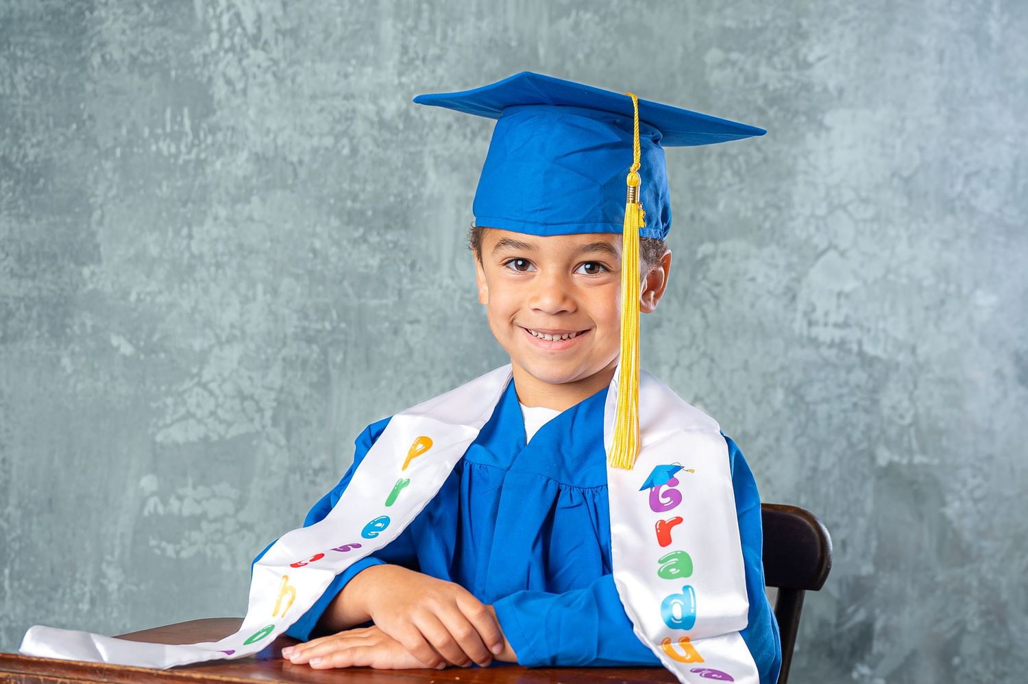 Young graduate in blue cap and gown sits on wooden chair against chalkboard background during preschool graduation photos.