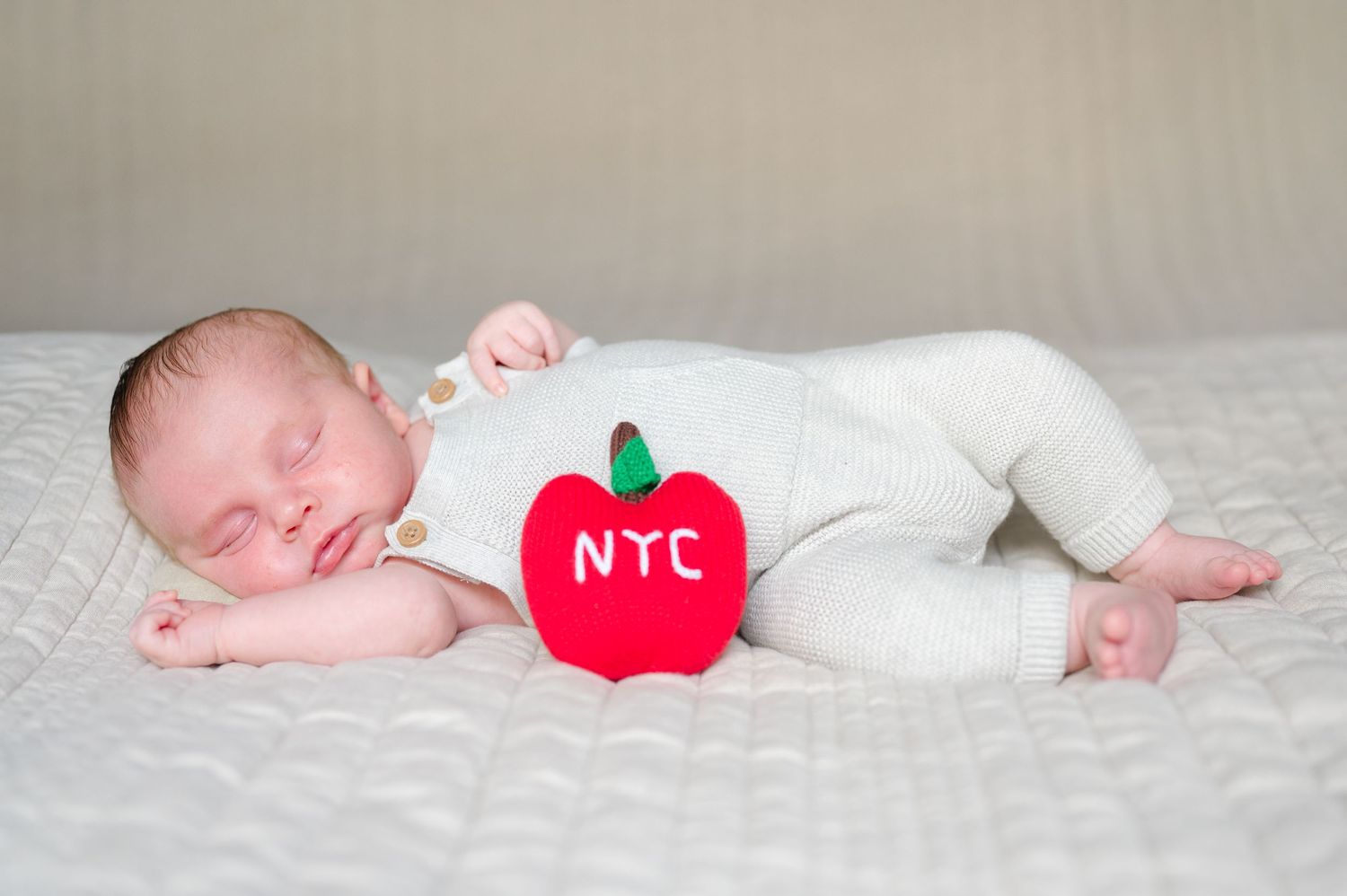 A sleeping newborn baby wearing white lies on soft fabric with a red apple decoration.
