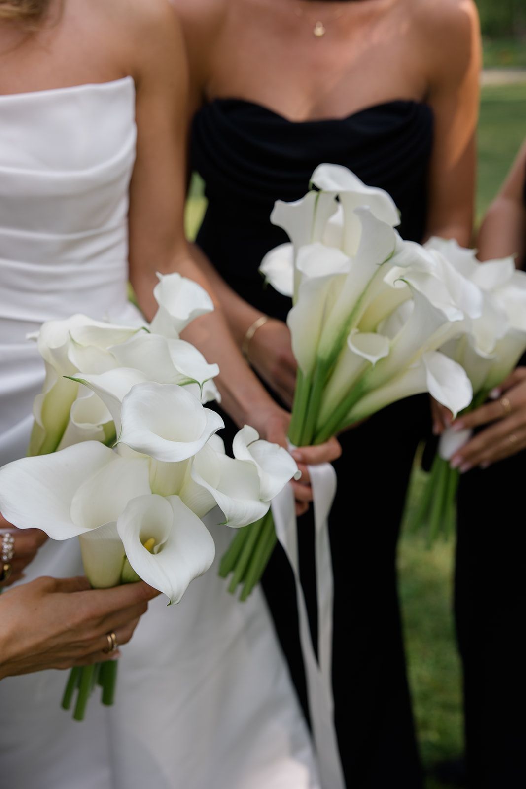 A row of bridesmaids in black dresses hold elegant white calla lily bouquets during an outdoor wedding ceremony.