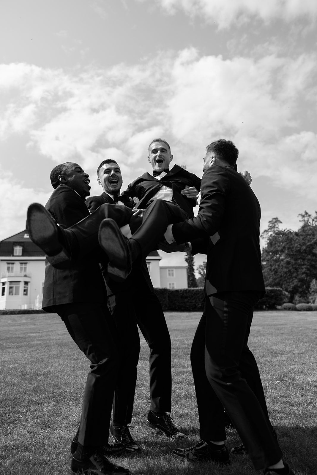 Black and white photo sequence shows groomsmen playfully wrestling and horsing around on a grassy lawn.