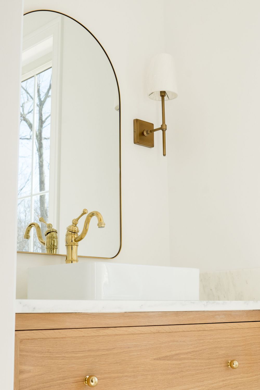 Bathroom vanity with curved mirror, brass fixtures, and light wood cabinetry against white walls.