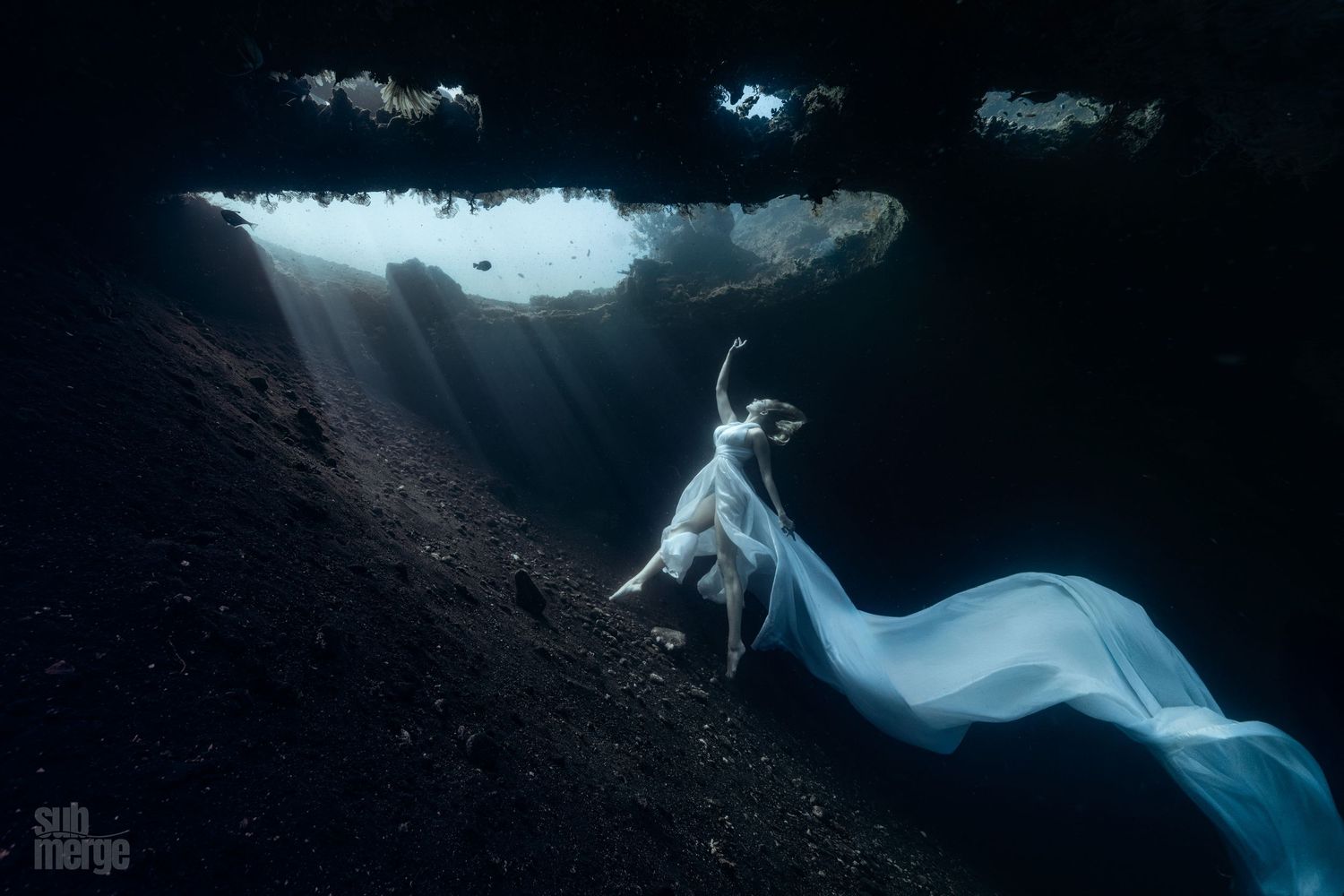 Underwater photographer captures ethereal images of a figure in flowing white fabric against dark coral formations.