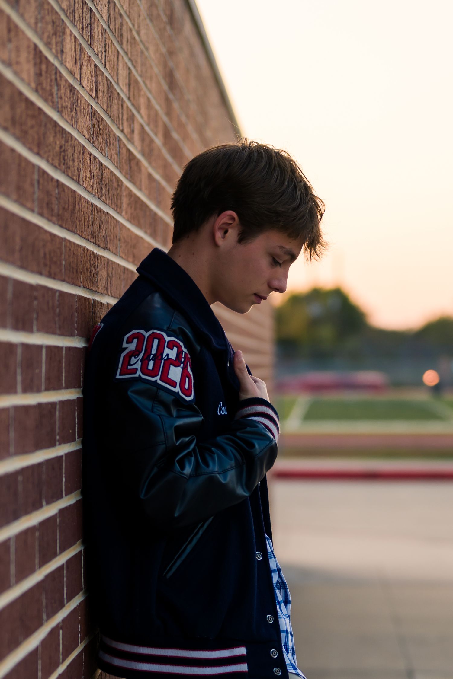 A student in a varsity jacket leans against a brick wall at sunset, creating a moody silhouette.
