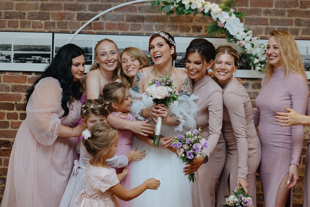 Wedding party in blush pink dresses poses together outdoors against brick wall with floral bouquets.