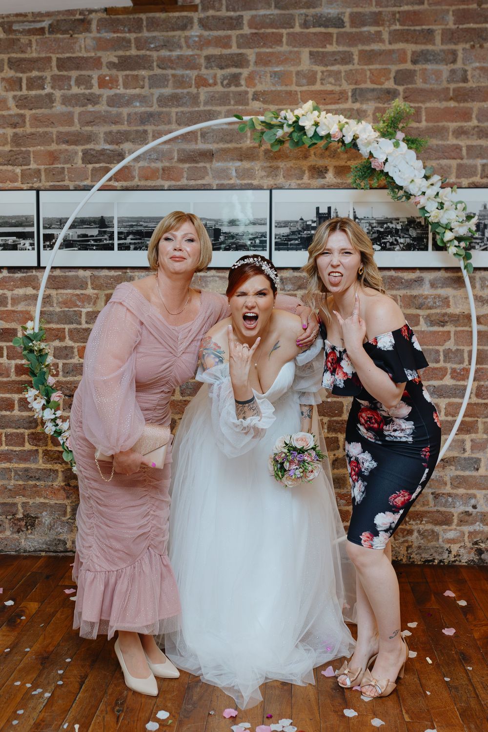 Wedding party members in pink dresses and white gown celebrate joyfully in front of exposed brick wall.