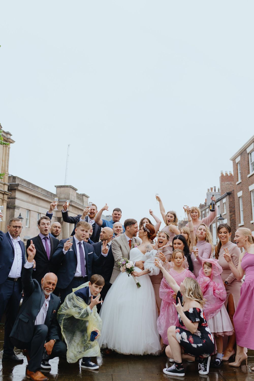 A large wedding party poses together on a rooftop, celebrating with raised arms and joyful expressions.