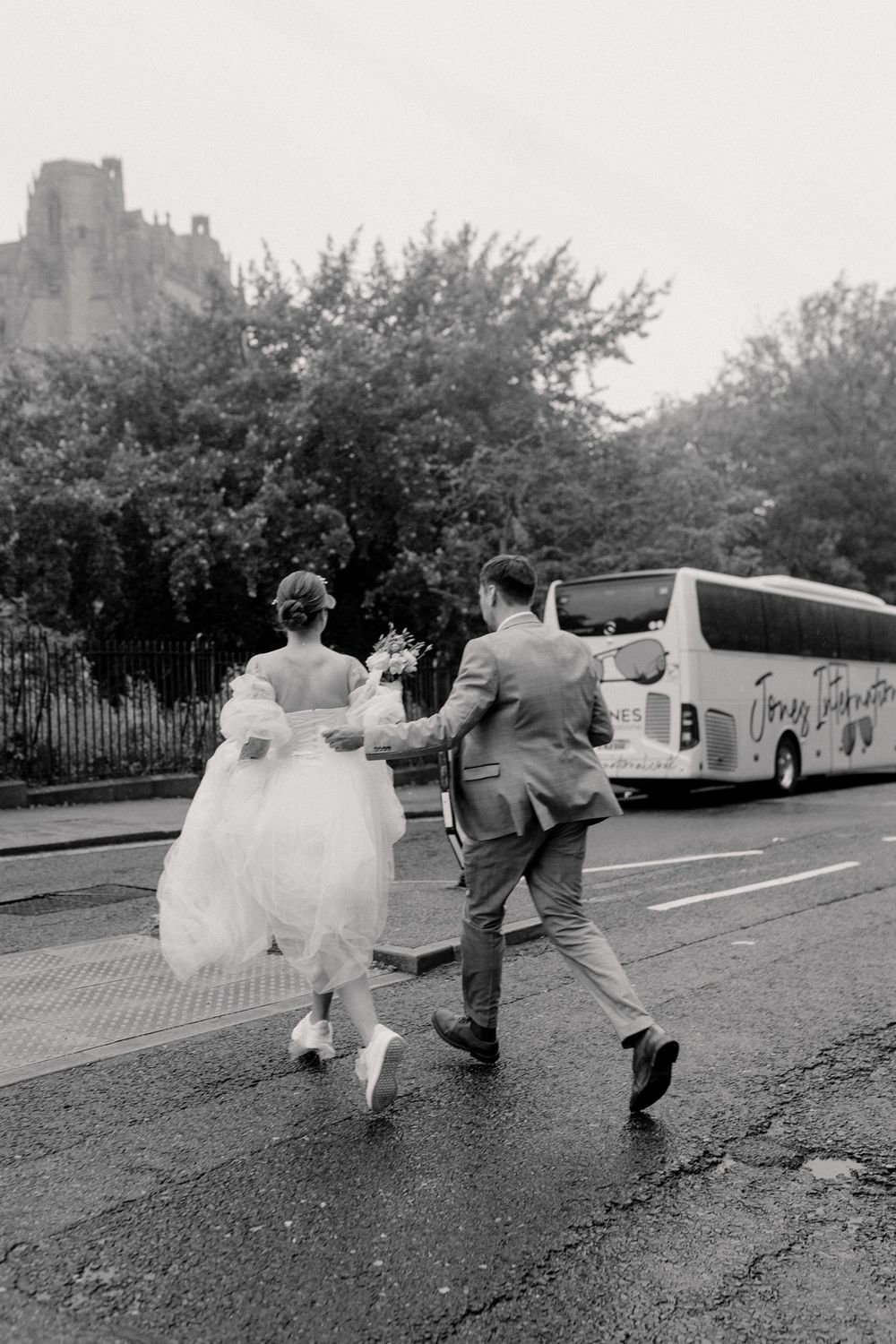 A couple in wedding attire runs down a street with a tour bus visible in the background.