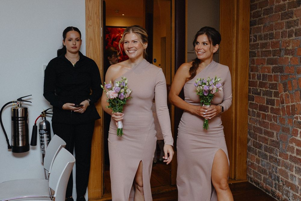 Wedding party in blush pink dresses walks through a brick-lined hallway holding small floral bouquets.