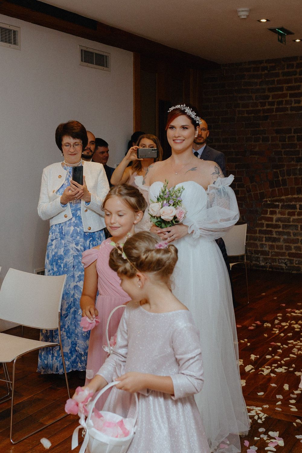 Wedding preparations show bridesmaids helping with final dress adjustments in a rustic brick-walled room.